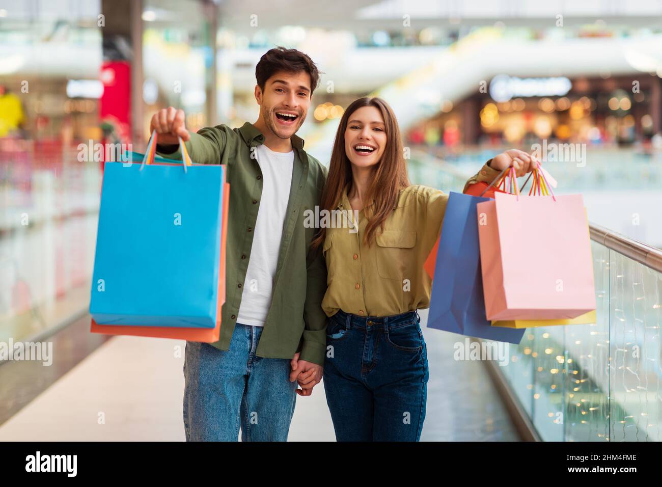 Cheerful Spouses Showing Shopping Bags To Camera Standing In Mall Stock Photo