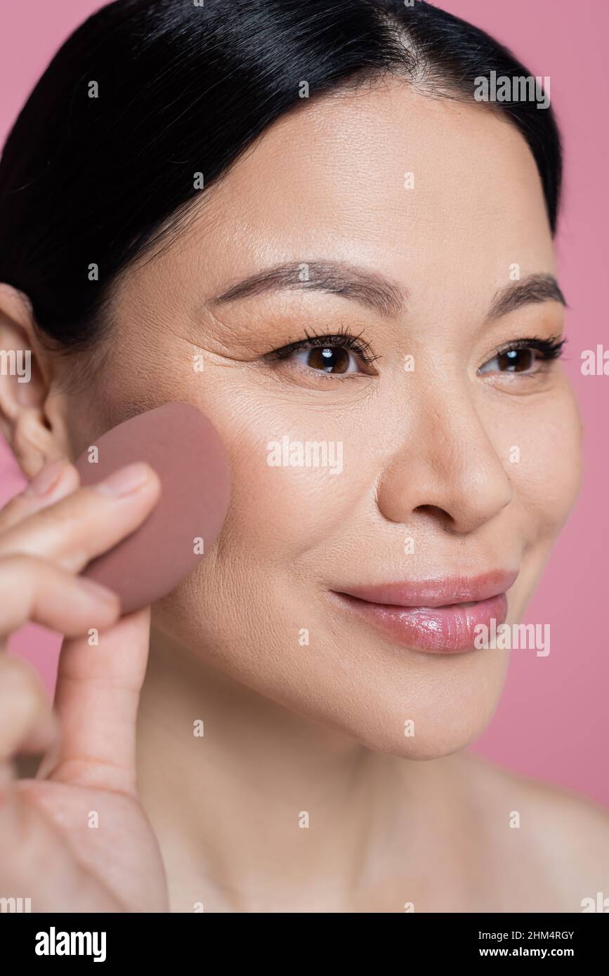 Close up view of asian woman applying face foundation with blurred beauty blender isolated on pink Stock Photo