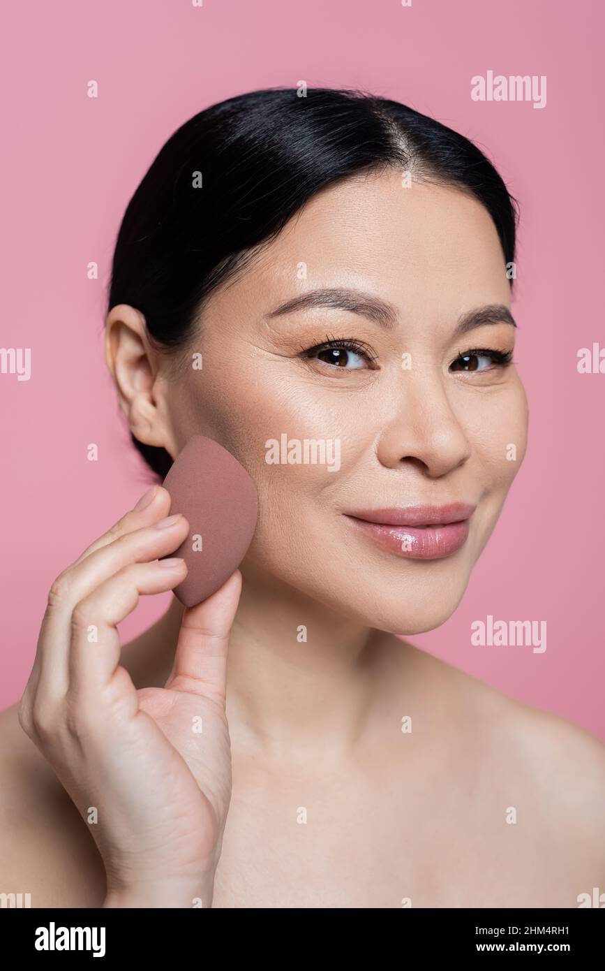 Smiling asian woman applying face foundation with beauty blender isolated on pink Stock Photo