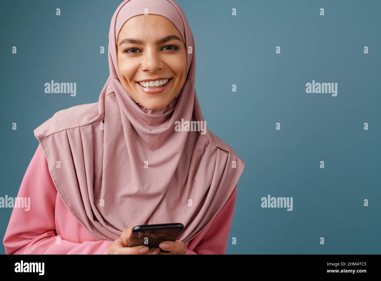 Young muslim woman in hijab smiling while using mobile phone isolated ...