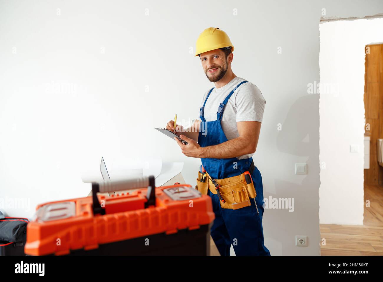 Smiling handyman worker, builder in uniform and hard hat looking at camera, making notes on a clipboard while standing indoors Stock Photo