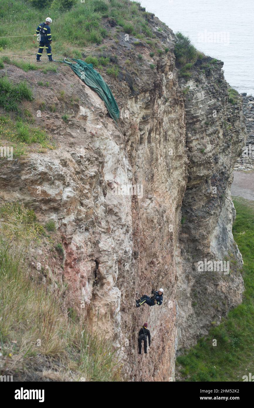 Long view of firemen on training exercise abseiling down cliff face in mock rescue of figure which in this case is a dummy Stock Photo
