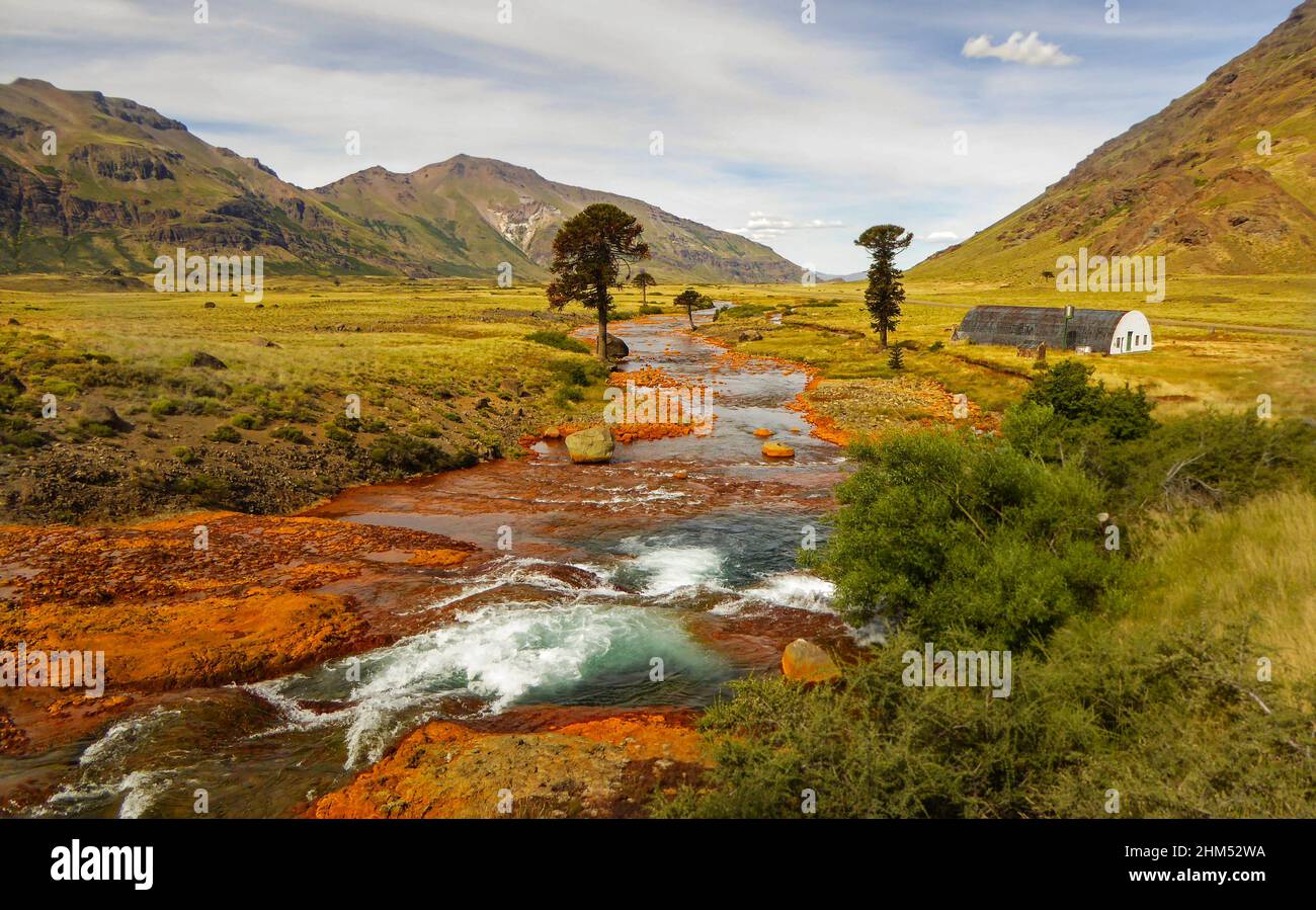 Bridge over a river with orange shores with mountains at the background and pehuen trees. Río Agrio, Caviahue, Neuquén, Argentina. Stock Photo