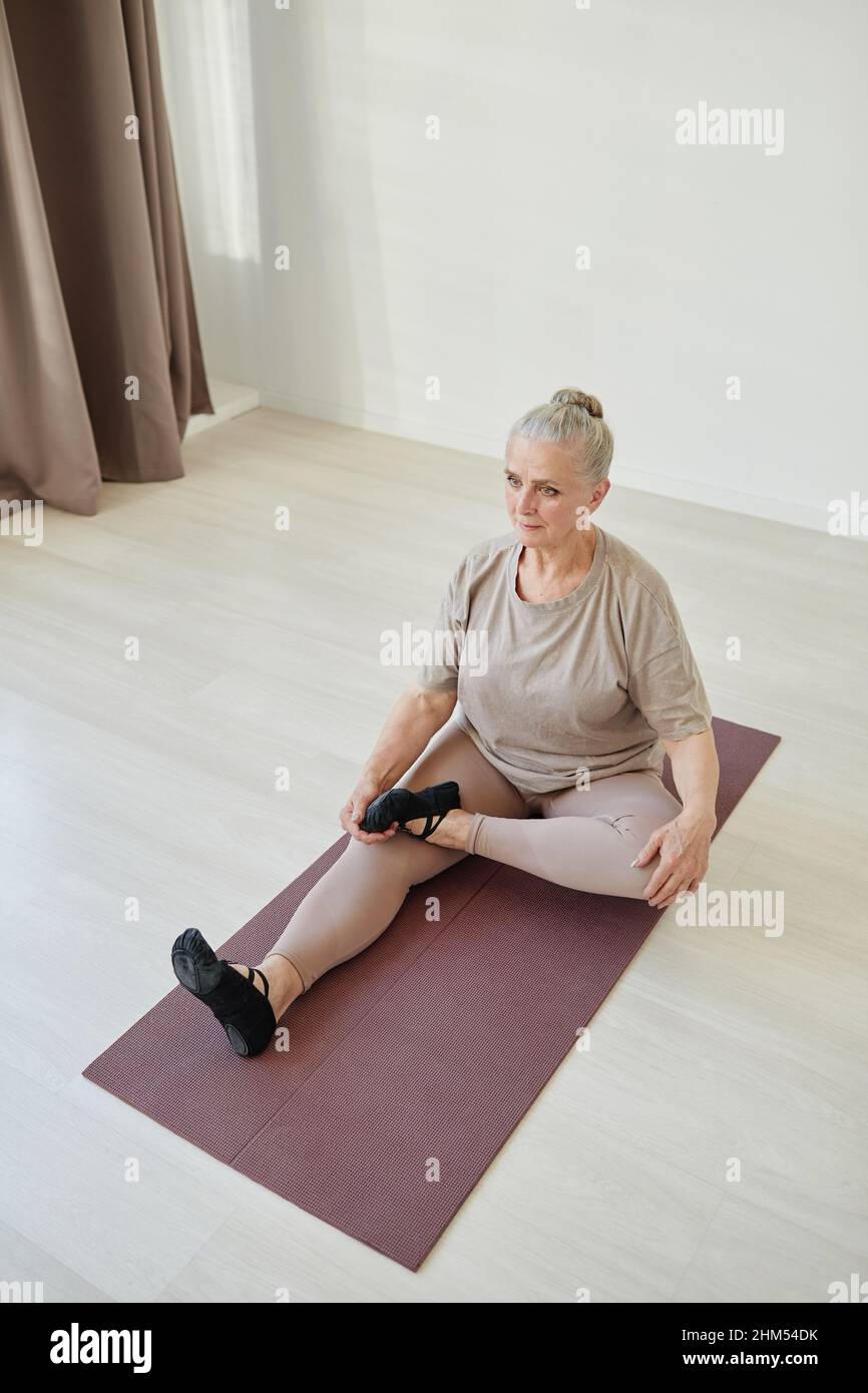 Senior active woman with grey hair sitting on mat and keeping left leg bent in knee on the right one during yoga exercise Stock Photo