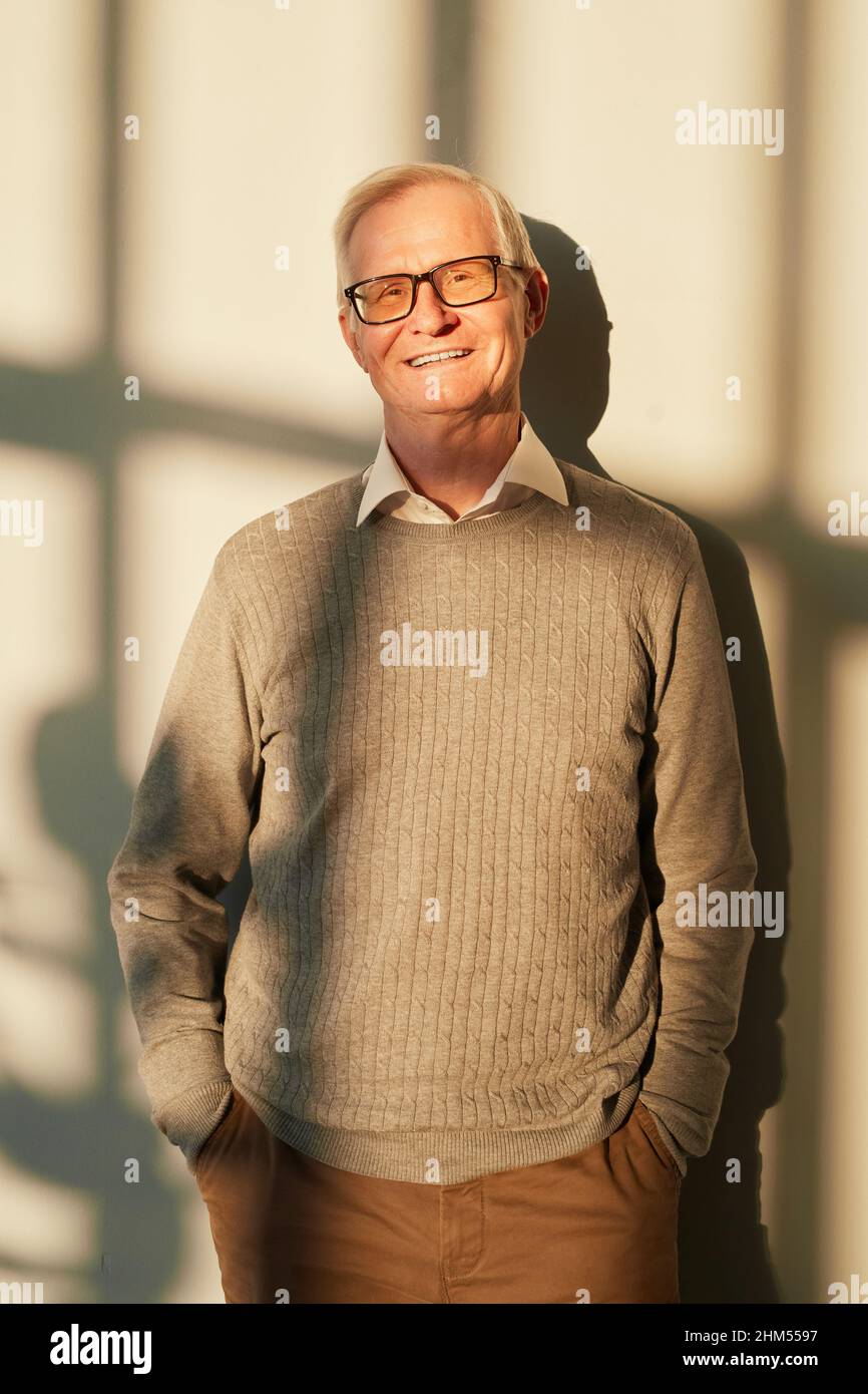 Cheerful senior man in beige pullover and brown pants standing sunlit against wall and looking at camera Stock Photo