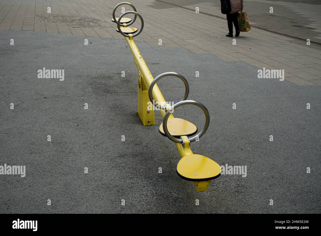 A children's seesaw swing on an children playground in a pedestrian zone. Someone walks by the playground. Stock Photo
