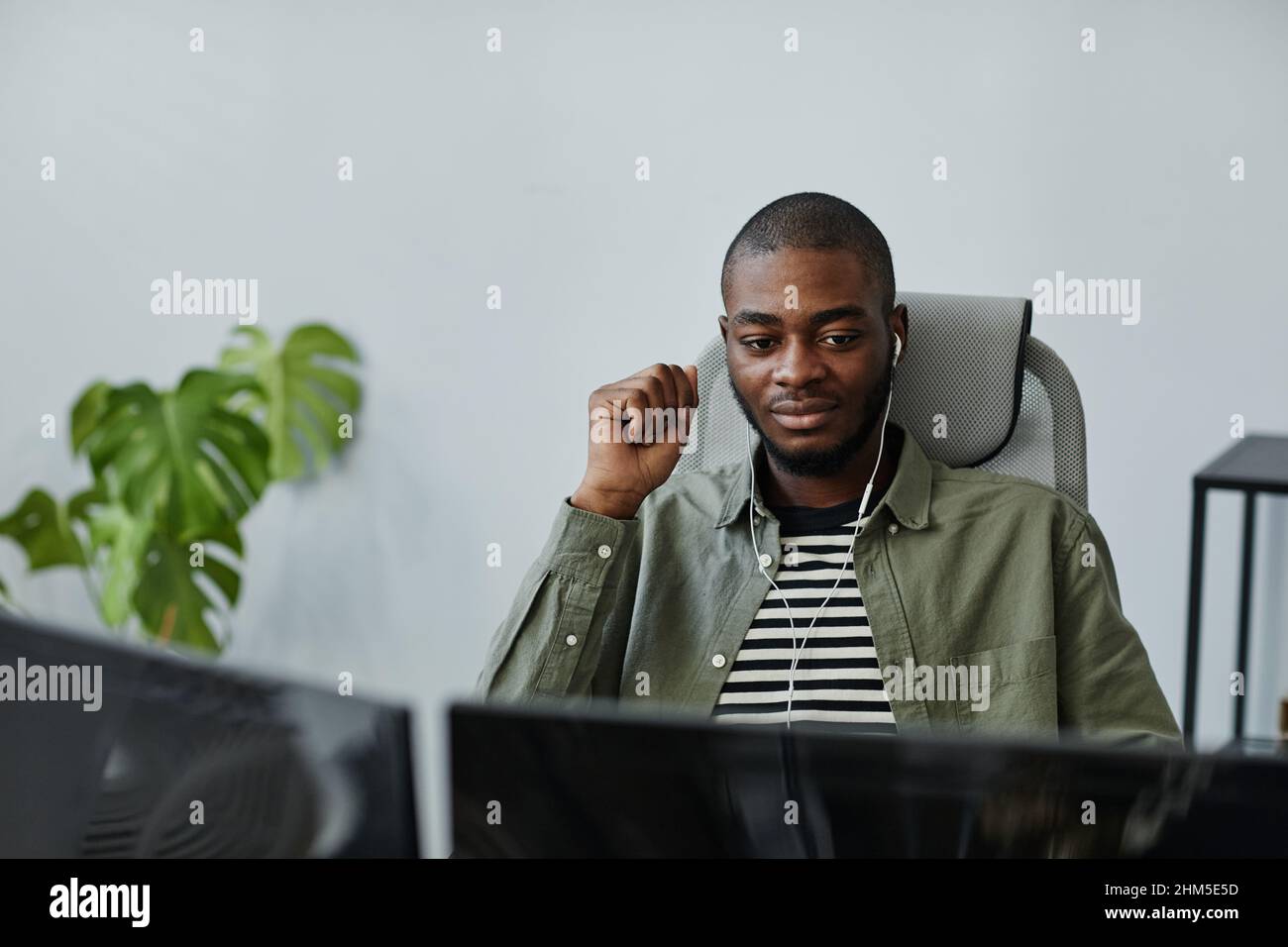 Young African American businessman in earphones and casual apparel working in the net while sitting in front of computer screen Stock Photo