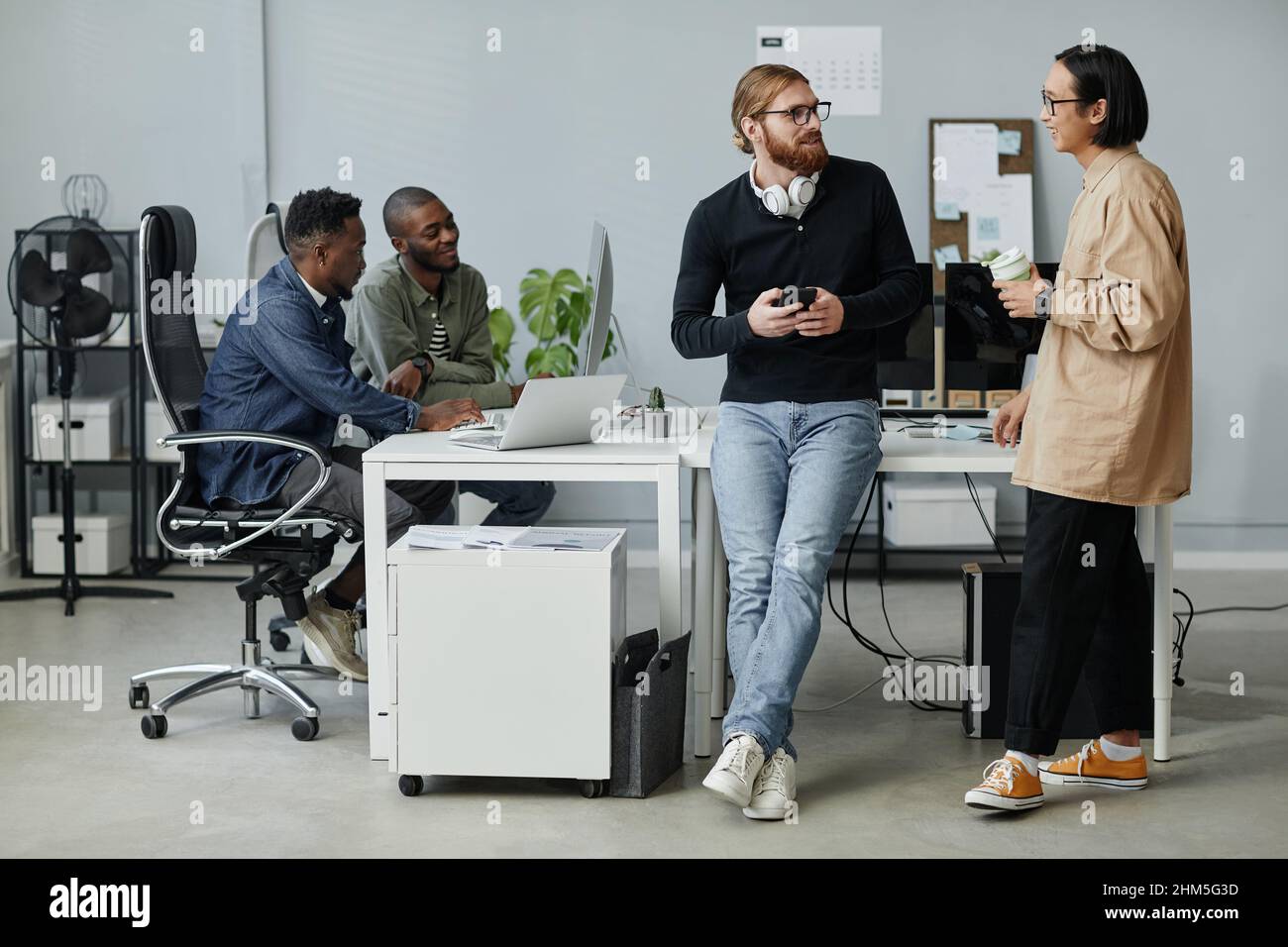 Young contemporary program managers talking at break while their two colleagues working in front of computer in office Stock Photo