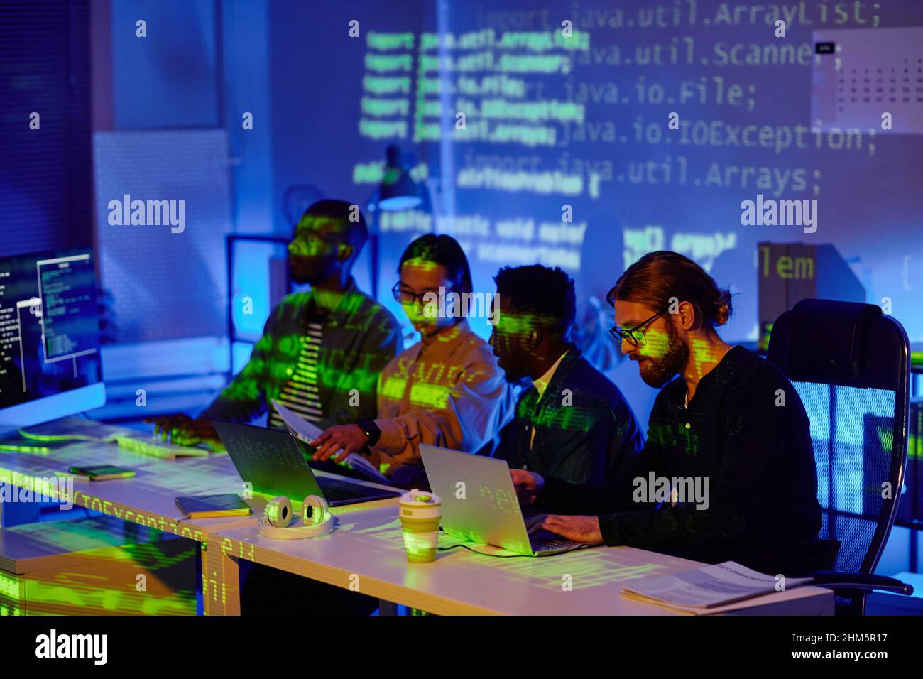 Several young contemporary software developers sitting in row by desks ...