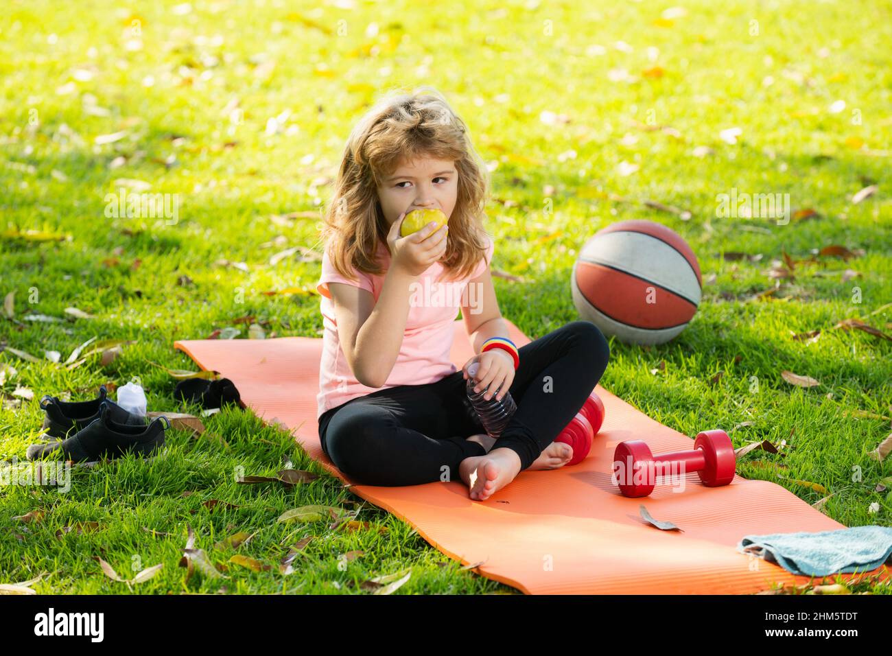 Healthy lifestyle and healthy food concept. Little boy child in sportswear eating apple sitting on sport mat after training on sunny spring day Stock Photo