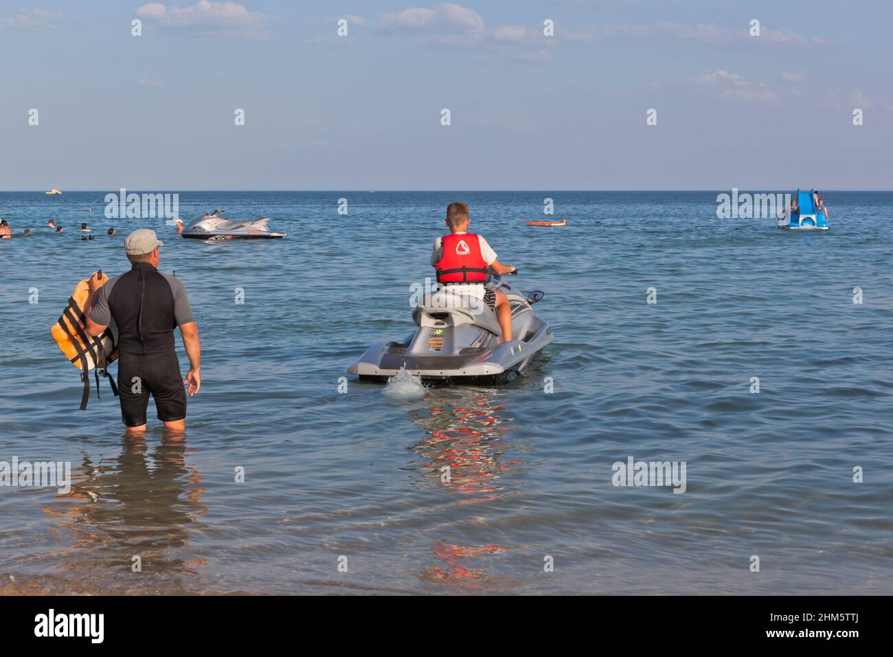 village Zaozernoe, Evpatoria, Crimea, Russia - July 18, 2021: A boy under the supervision of an instructor drives off on a jet ski from the Super Aqua Stock Photo