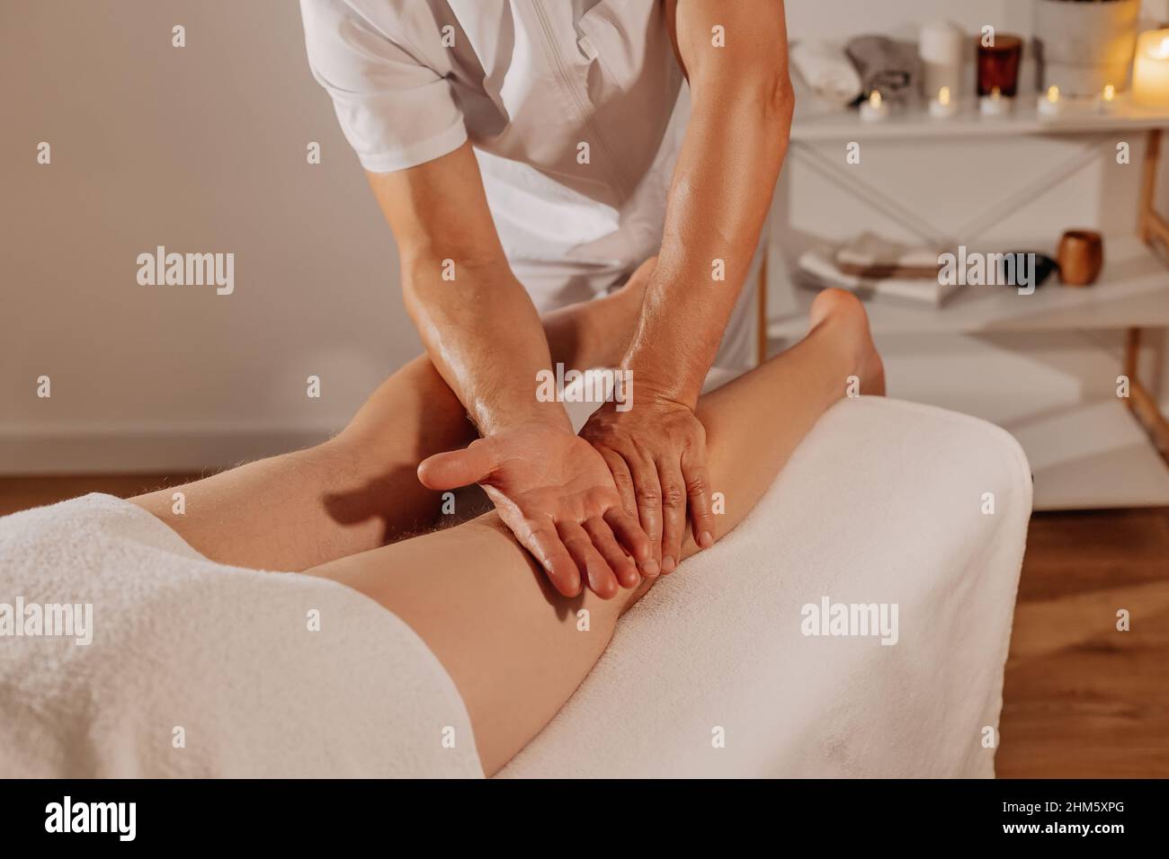 Professional legs massage in SPA salon on the background of candles. Handsome masseur therapist in white uniform making manual therapy for athlete muscles. Concept of wellness, body and health care. Stock Photo