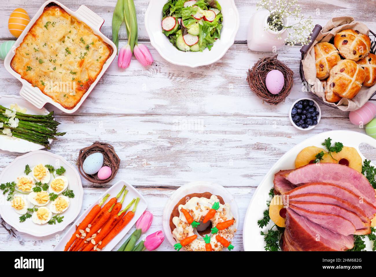 Classic Easter ham dinner. Above view frame on a white wood background with copy space. Ham, scalloped potatoes, vegetables, eggs, hot cross buns and Stock Photo