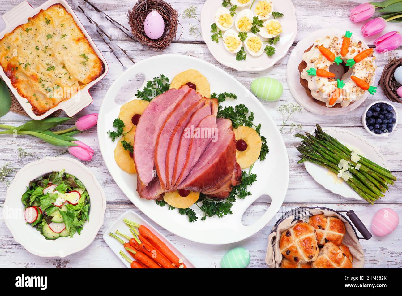 Traditional Easter ham dinner. Top view table scene on a white wood background. Ham, scalloped potatoes, eggs, hot cross buns, carrot cake and vegetab Stock Photo