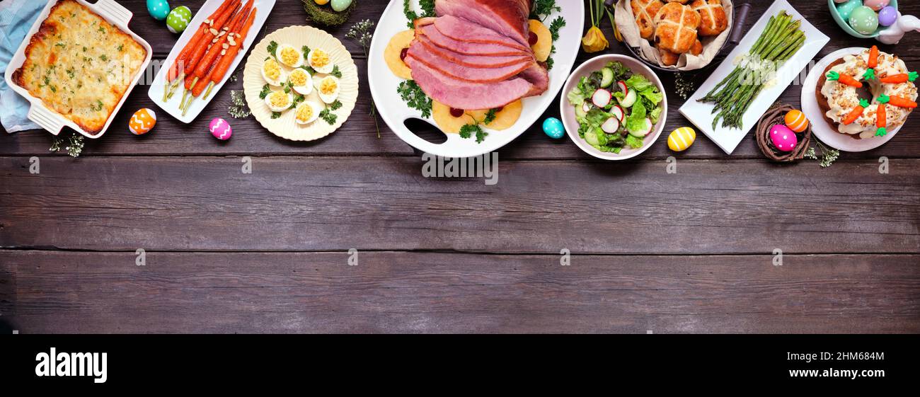 Traditional Easter ham dinner. Above view top on a dark wood banner background with copy space. Ham, scalloped potatoes, vegetables, eggs, hot cross b Stock Photo