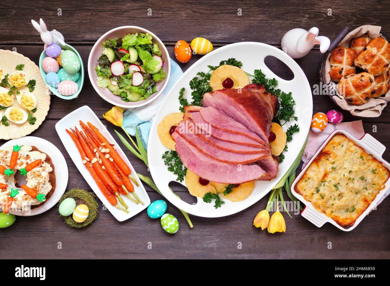 Traditional Easter ham dinner. Top down view table scene on a dark wood background. Ham, scalloped potatoes, vegetables, eggs, hot cross buns and carr Stock Photo