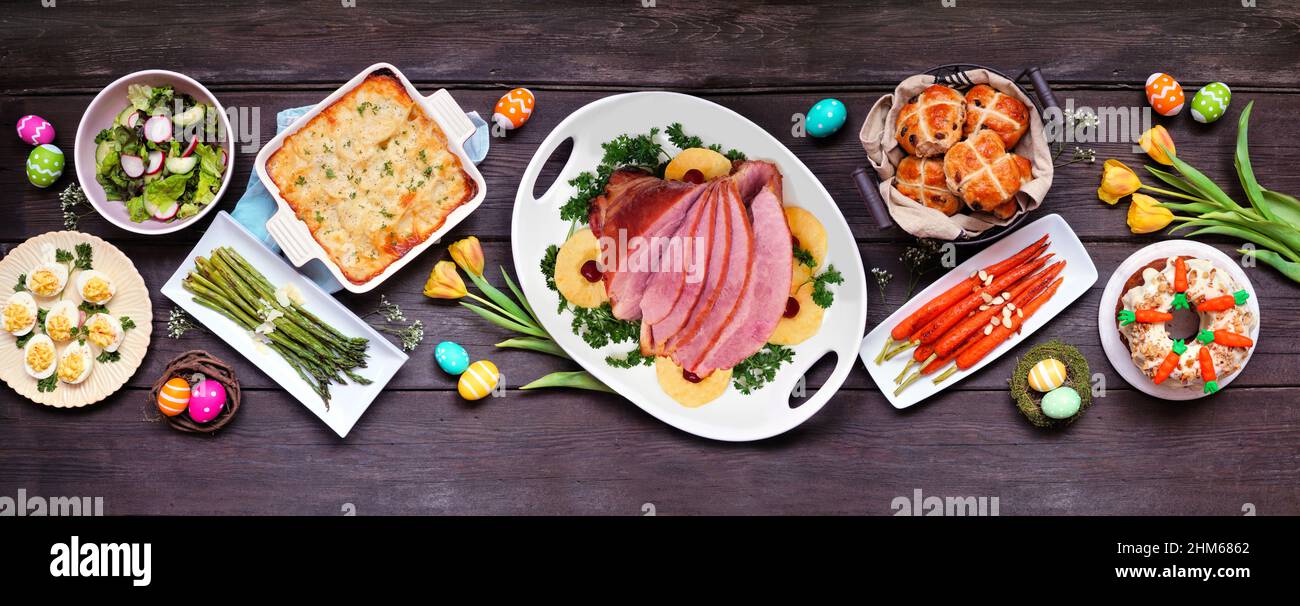 Classic Easter ham dinner. Top view table scene on a dark wood banner background. Ham, scalloped potatoes, eggs, hot cross buns, carrot cake and veget Stock Photo