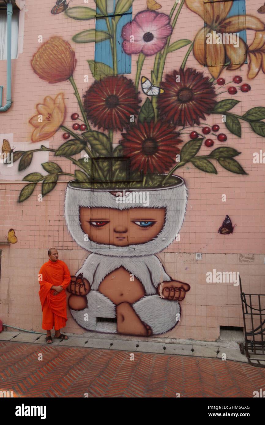 Buddhist Monk standing by a painting by Thai artist Alex Face featuring his famous character Mardi; Klong Ong Ang Walking Street, Bangkok, Thailand Stock Photo