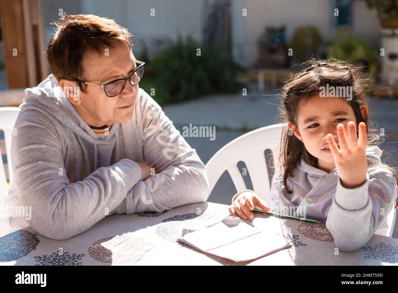 Beautiful girl doing homework with the help of her grandmother on an outdoor patio.Close up portrait of senior teacher supervising little kid doing homework back yard. Young child counting on fingers. Stock Photo