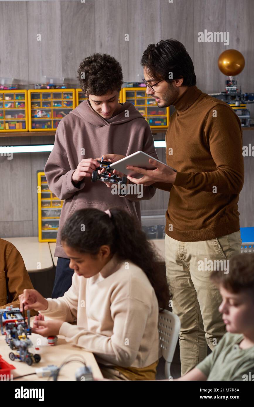 Vertical portrait of young male teacher helping diverse group of children building robots in engineering class at school Stock Photo