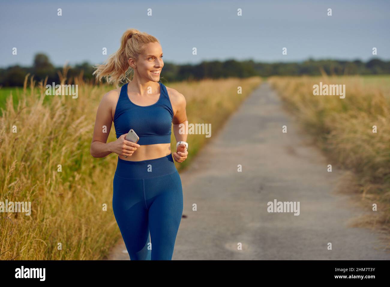 Fit healthy young woman enjoying a jog along a country road passing the camera with a happy smile full of vitality in an active lifestyle concept Stock Photo