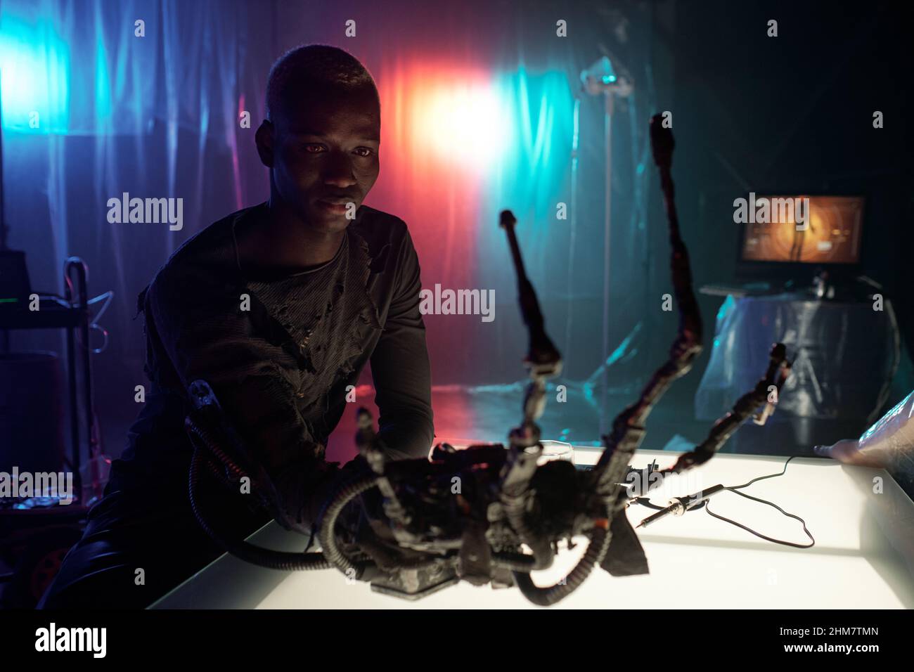 African cyberpunk man sitting at the table and examining his cyber arm in dark room with neon lights Stock Photo