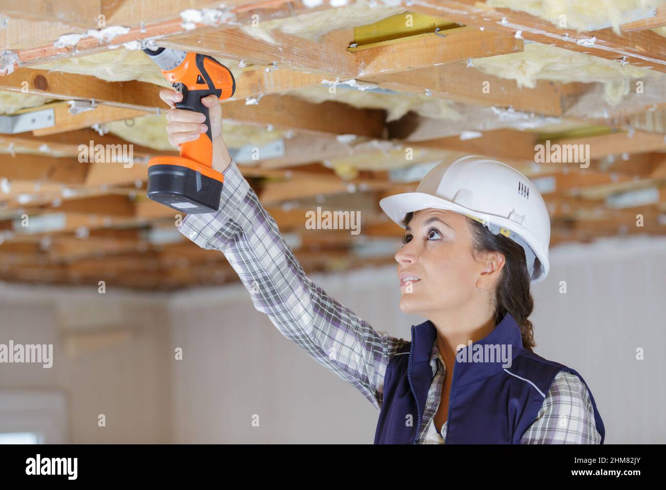female builder using cordless drill on wooden ceiling joist Stock Photo