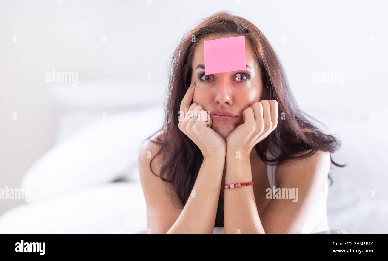 Woman who forgot something has pink postit on her forehead holding her head with both heads, thinking hard. Stock Photo