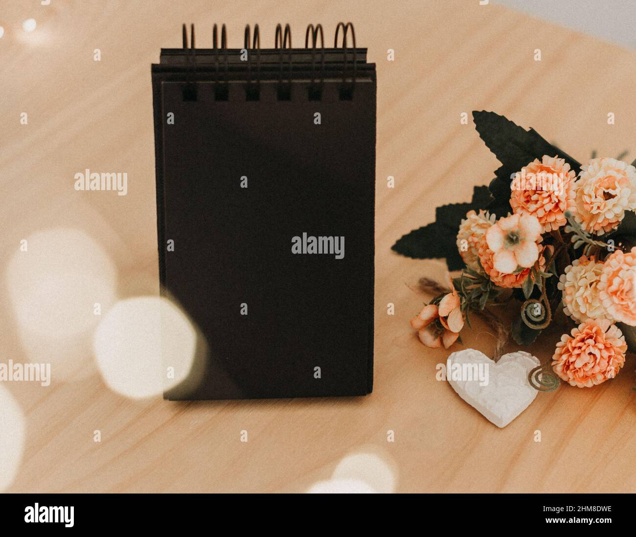 notebook with black sheets with copy space, on a wooden table with a bouquet of flowers and a white heart and bokeh lights around it. Stock Photo