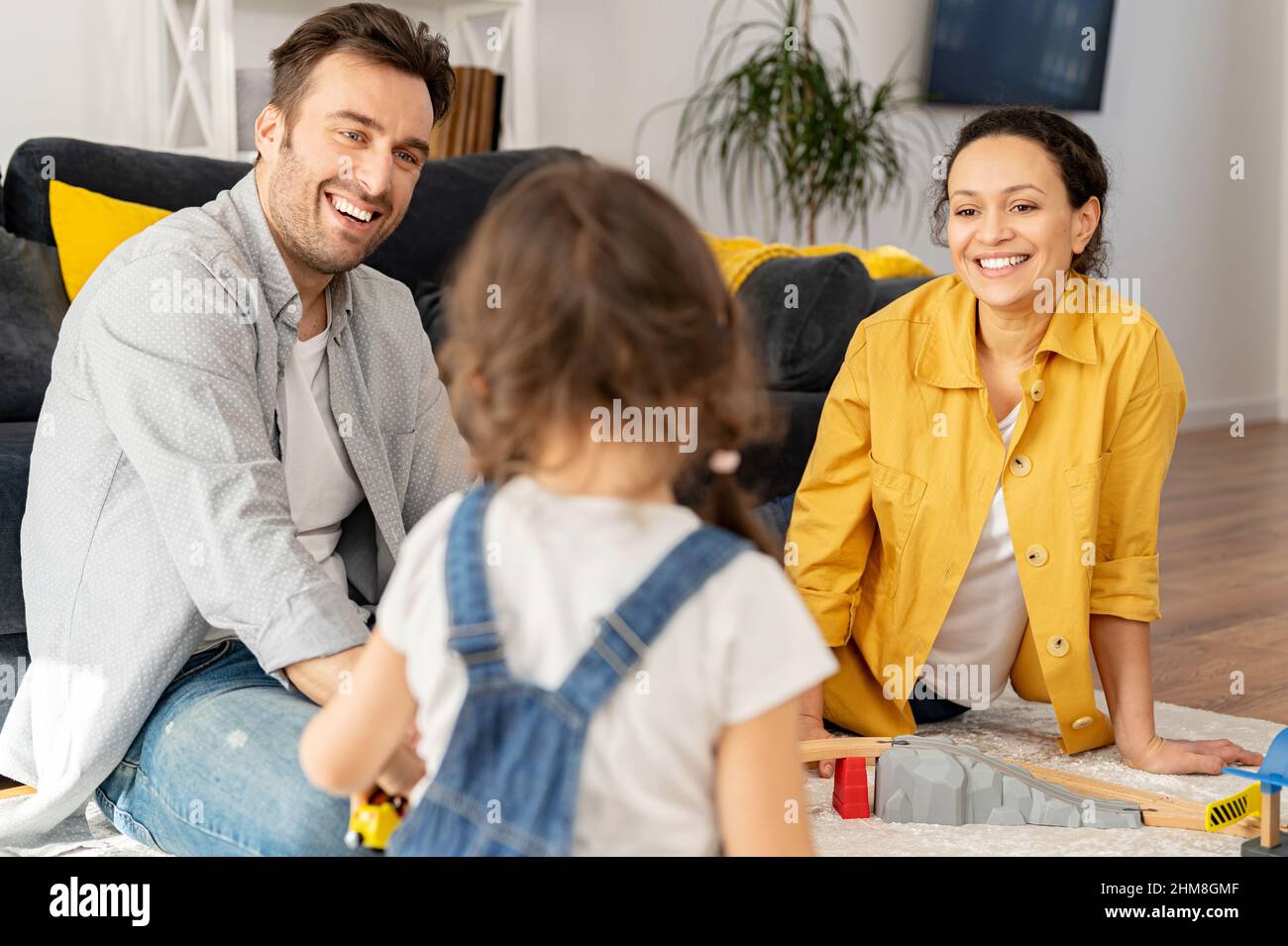 Love and care concept. Two parents looking at the little toddler daughter playing on the floor sitting back to camera, happy mother and father spending time together with kid Stock Photo