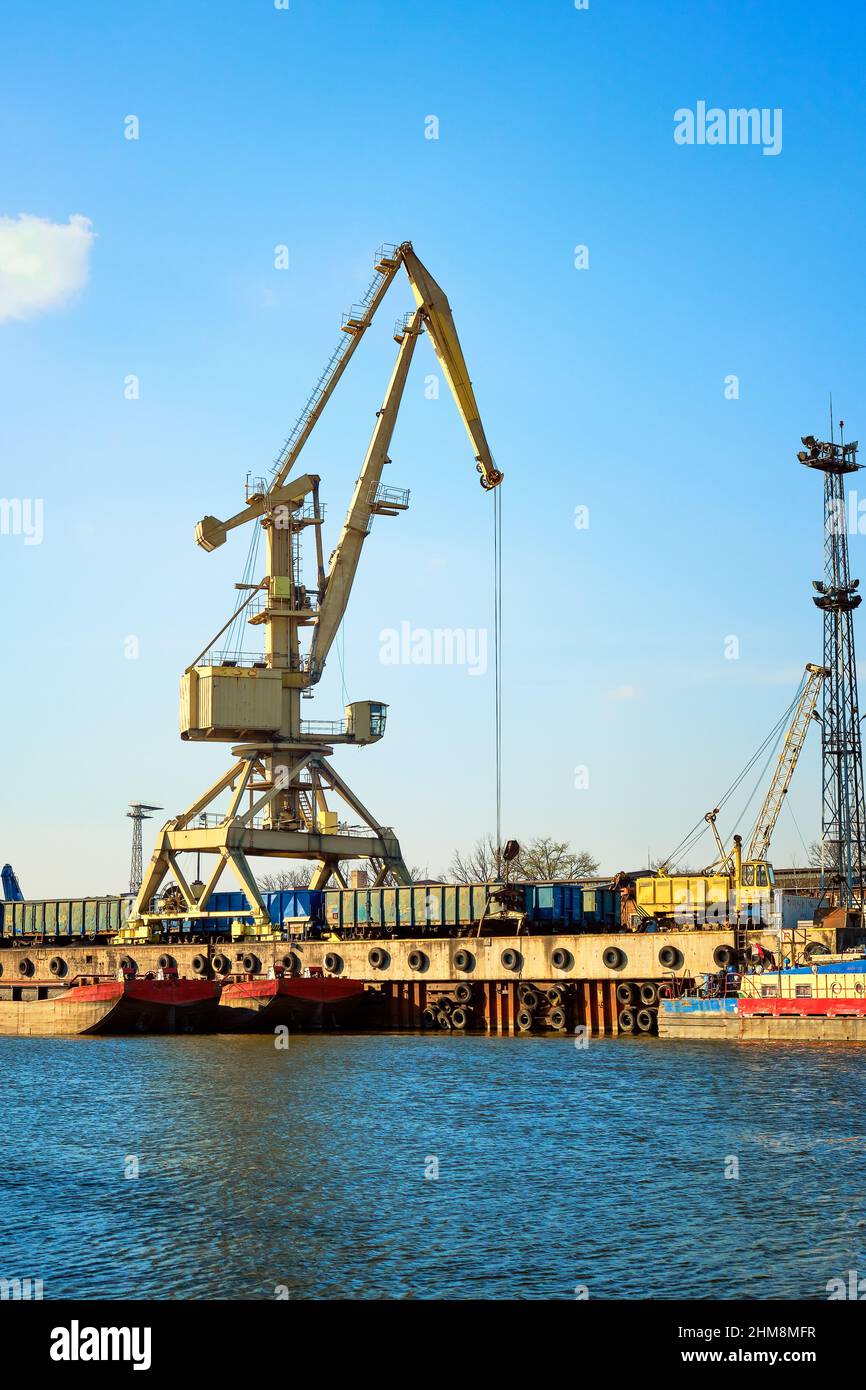 River port crane loading open-top gondola cars on sunny day. Empty river drag boats or barges by pier. Empty cars ready for loading. Vertical image Stock Photo