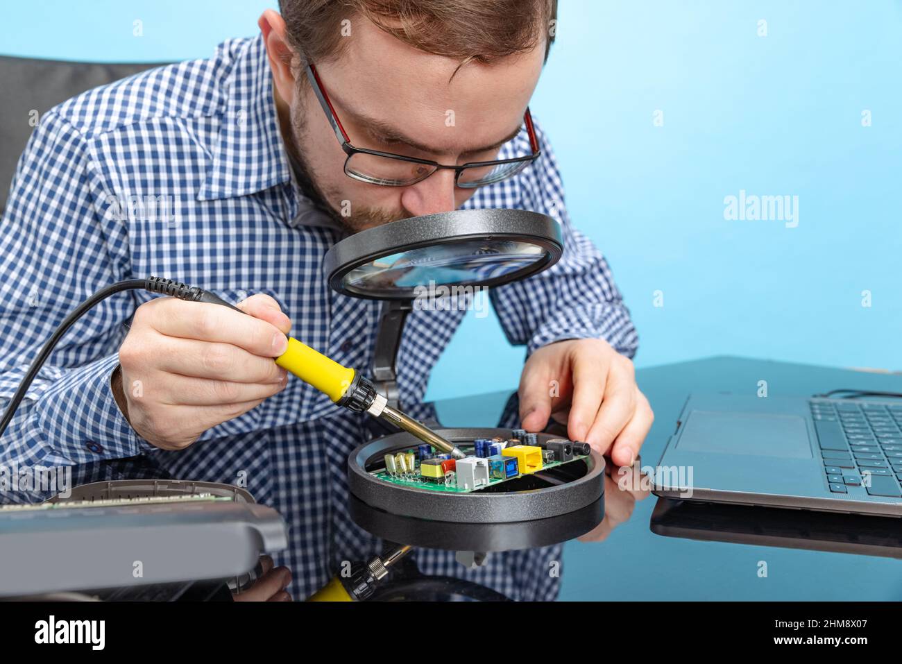 Portrait of concentrated man, computer geek looking in magnifying glass on motherboard isolated over blue background Stock Photo