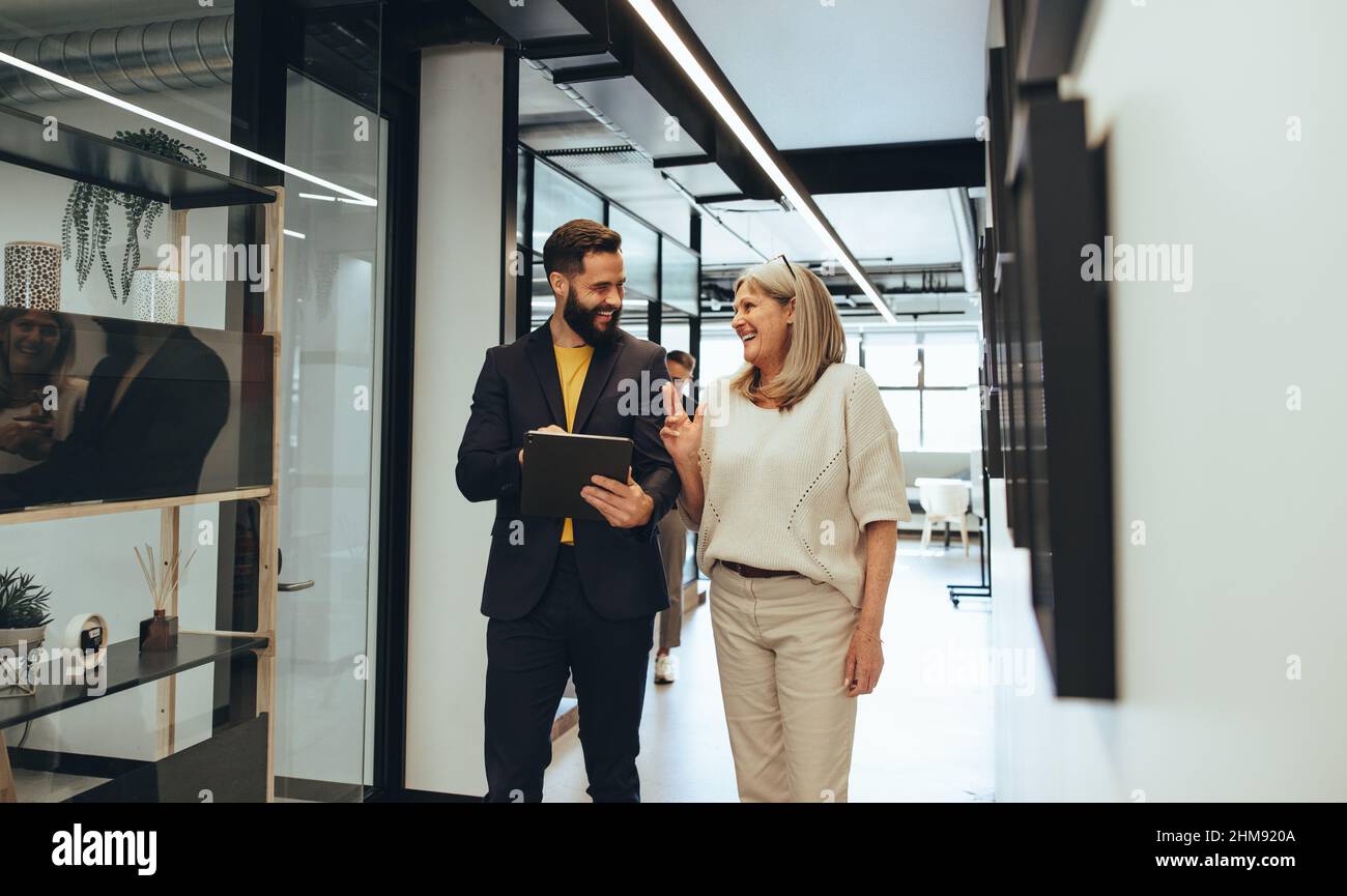 Diverse businesspeople sharing a laugh during a discussion in an office. Two happy business colleagues using a digital tablet while walking together i Stock Photo