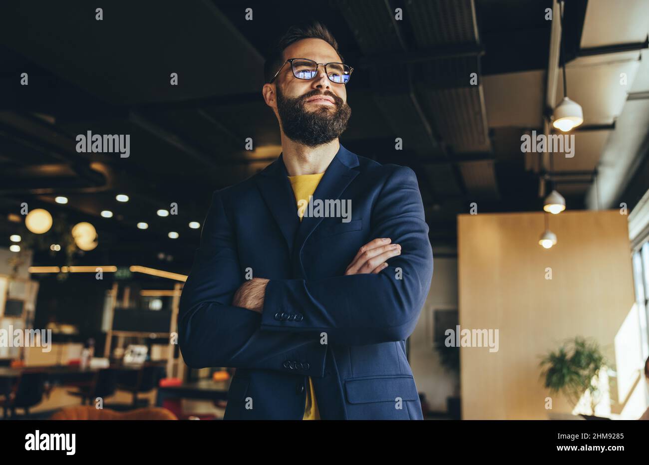 Cross-armed businessman looking away thoughtfully. Contemplative businessman thinking of new business ideas while standing in a co-working space. Youn Stock Photo