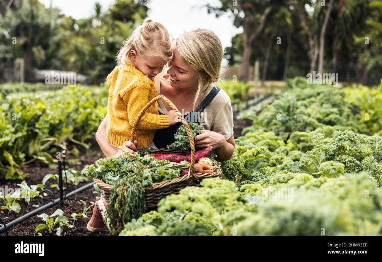 Bonding in the garden. Happy young mother smiling cheerfully while carrying her daughter and picking fresh kale in a vegetable garden. Self-sufficient Stock Photo