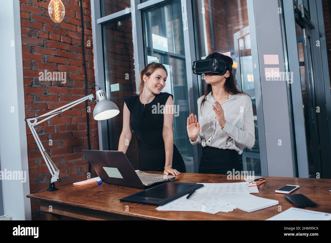 Two female application developers testing a new app designed for VR headset standing in modern office. Stock Photo