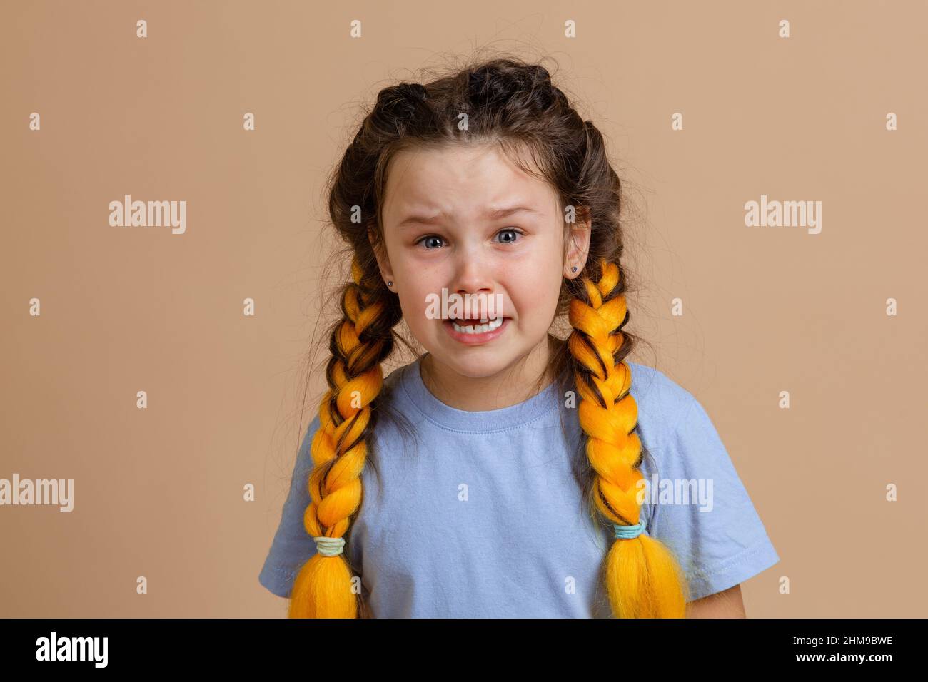 Sad Caucasian young girl crying with open mouth and wet eyes with kanekalon braids of yellow color on head wearing light blue t-shirt on beige Stock Photo