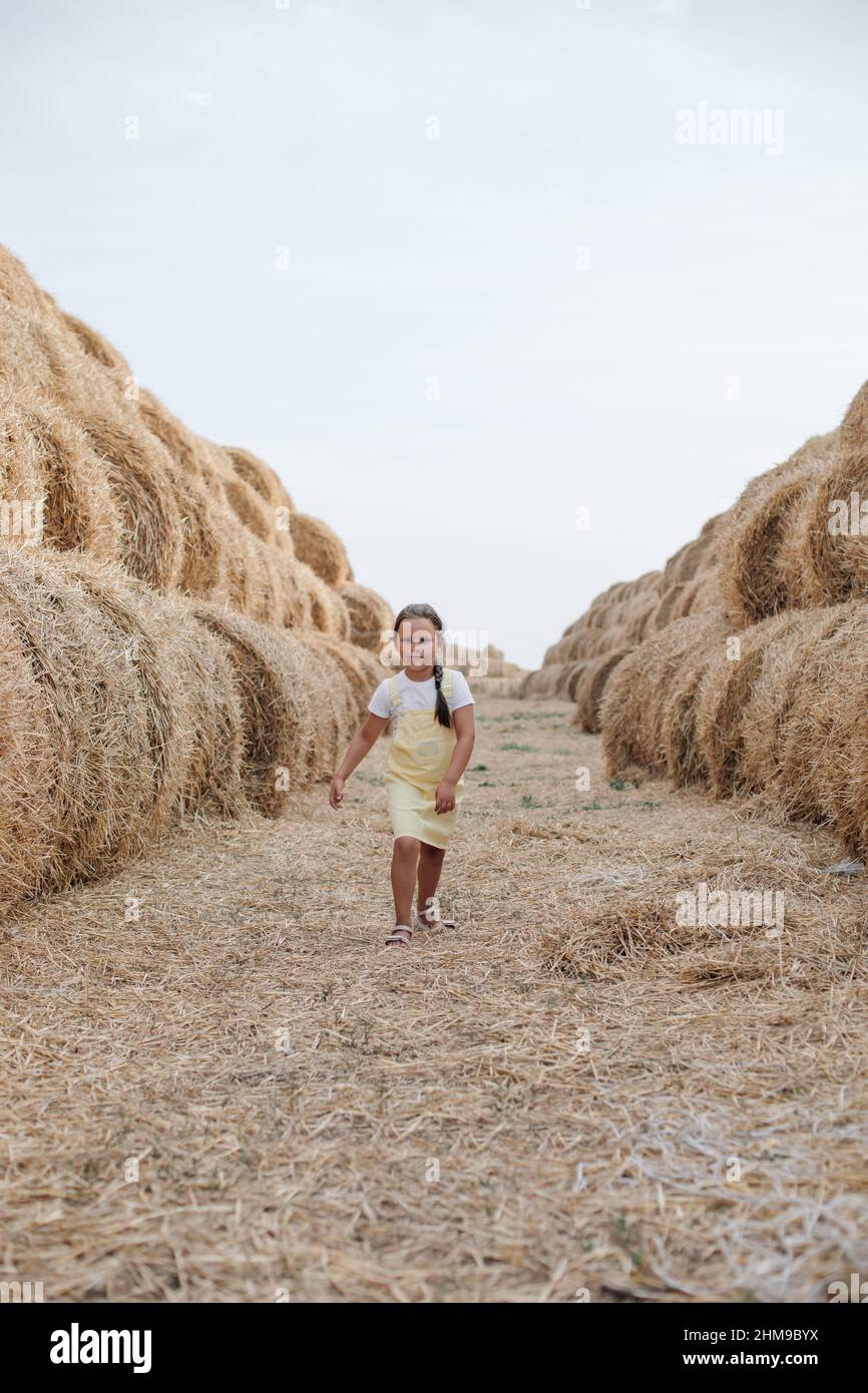 Portrait of young girl running between haystacks on covered with hay field country road looking ahead in sundress. Having fun away from city on field Stock Photo