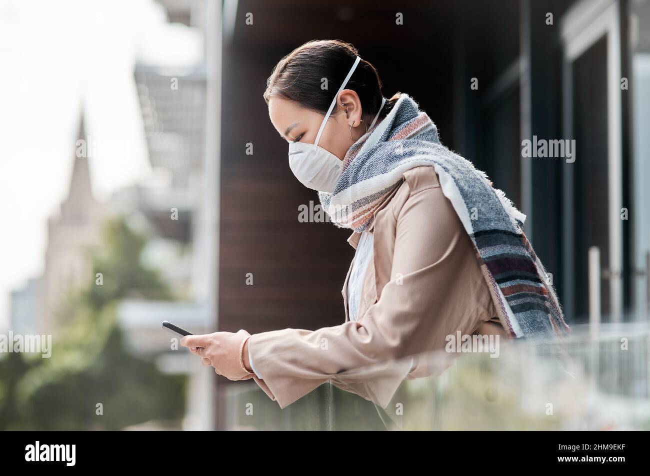 Technology allows us to be social while practising social distancing. Shot of a young woman wearing a mask and using a smartphone on the balcony at Stock Photo