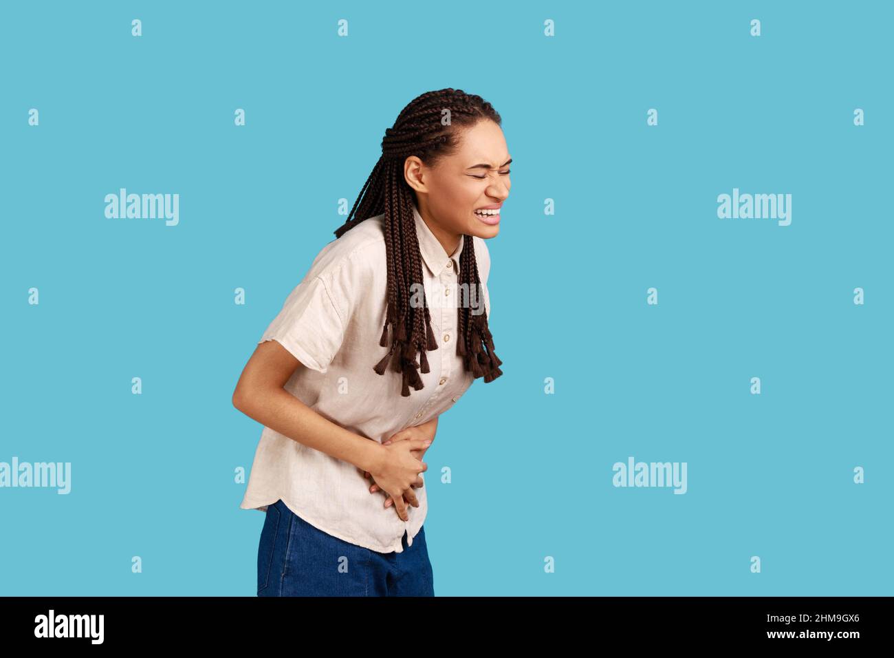 Displeased woman with black dreadlocks feels discomfort in stomach, keeps palms on tummy, has period cramps, ate spoiled food, wearing white shirt. Indoor studio shot isolated on blue background. Stock Photo