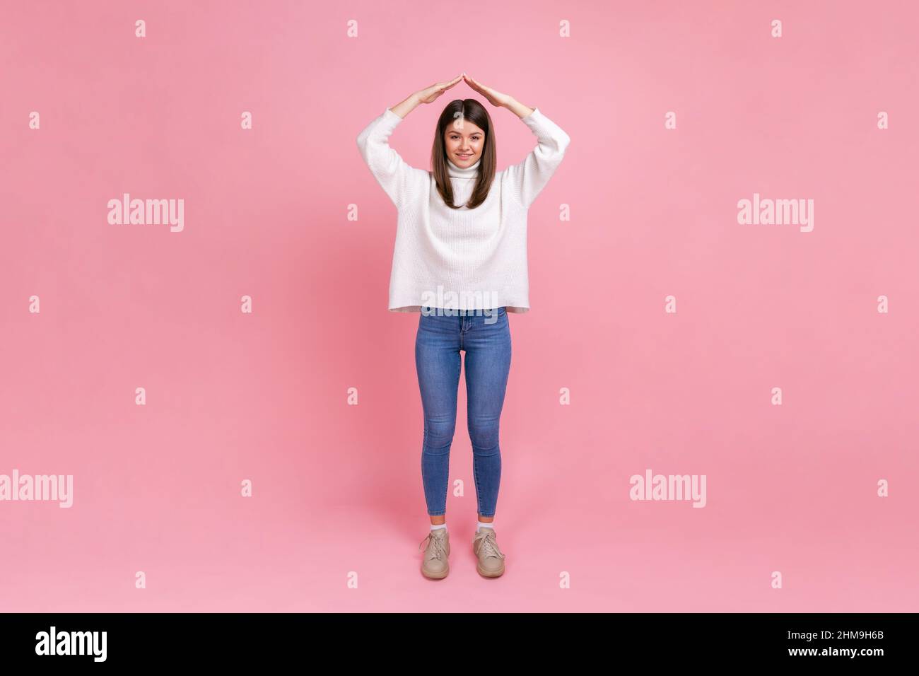 Full length portrait of attractive woman making roof of hands above head, feeling herself in safety, wearing white casual style sweater. Indoor studio shot isolated on pink background. Stock Photo