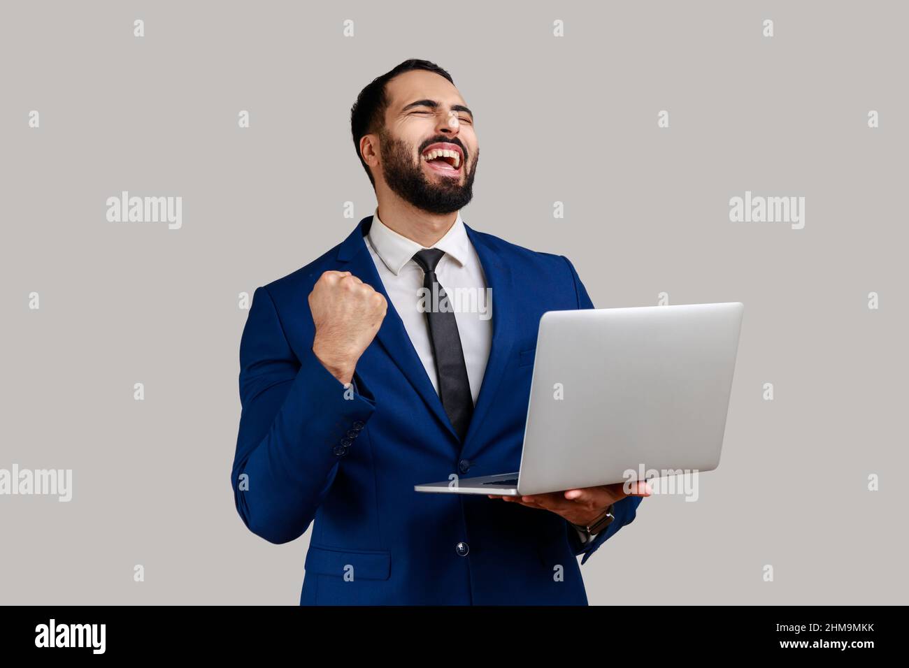 Portrait of excited bearded businessman screaming with joy and holding laptop, rejoicing victory, online betting, wearing official style suit. Indoor studio shot isolated on gray background. Stock Photo