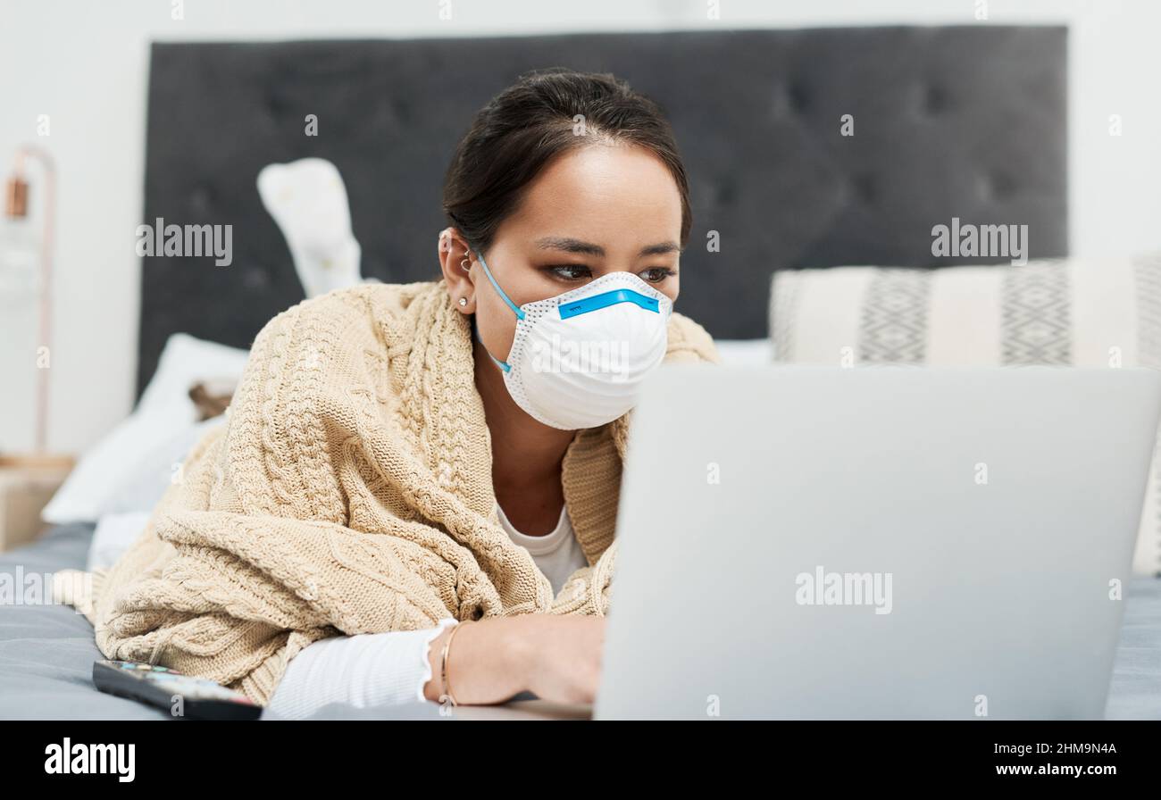 Keeping up with all the latest health advice. Shot of a woman wearing a mask while lying on her bed with her laptop. Stock Photo