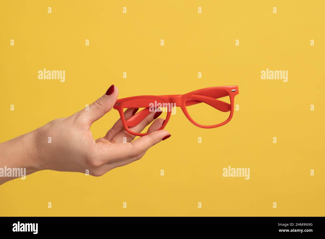 Profile side view closeup of woman hand holding and showing red eyeglasses frame. Indoor studio shot isolated on yellow background. Stock Photo