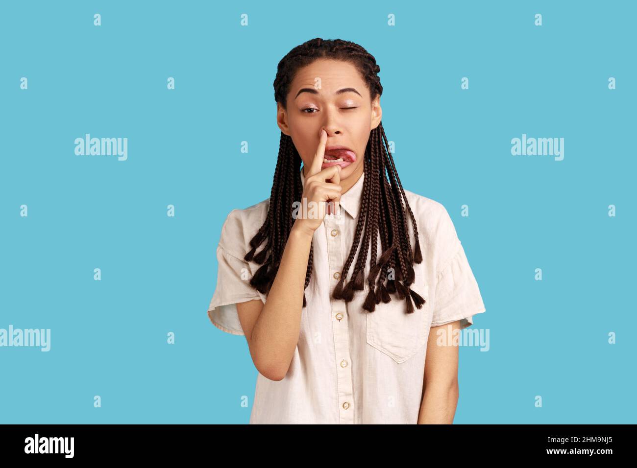 Portrait of funny impolite woman with black dreadlocks making crazy face with tongue out and picking her nose, bad manners, wearing white shirt. Indoor studio shot isolated on blue background. Stock Photo