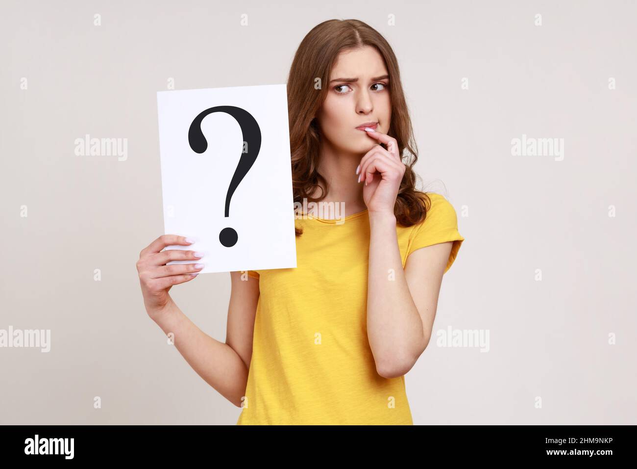 Young pensive woman in casual style attire holding paper with question mark over, thoughtful, face thinking about question, very confused idea. Indoor studio shot isolated on gray background. Stock Photo