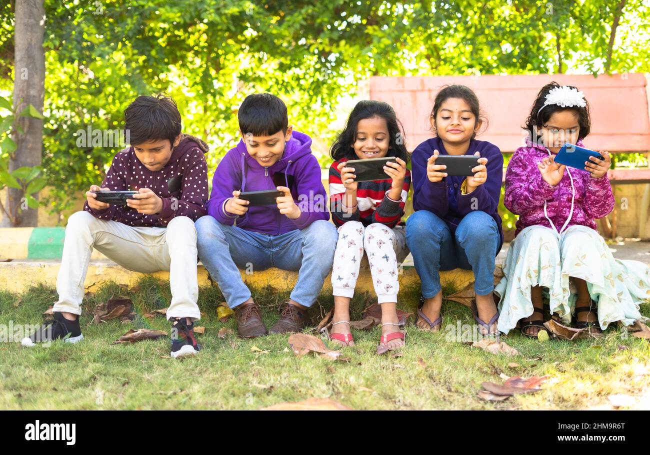 Group of kids busy using mobile phone while sitting at park - concept of smartphone addiction, playing video game, reduced physical activity and Stock Photo