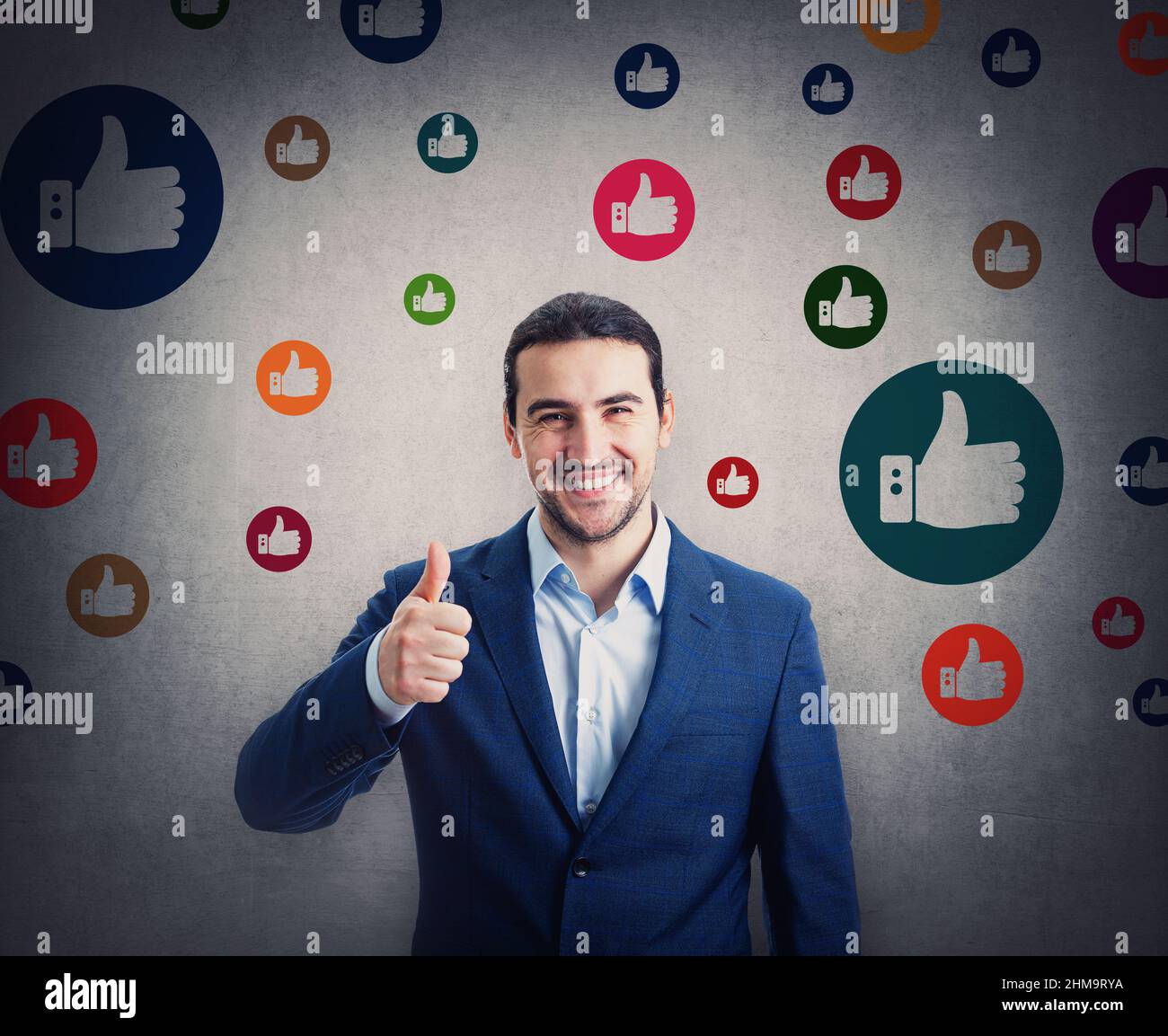Contented businessman smiling broadly shows thumb up gesture, surrounded by multicolor like symbols. Business project approval and positive feedback Stock Photo
