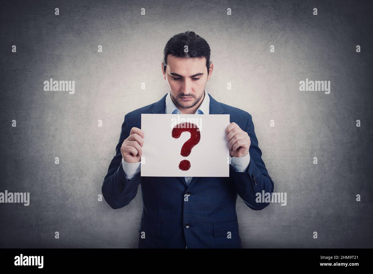 Doubtful businessman holding a paper sheet with question symbol, isolated on grey wall background. Depressed person looking down pensive. Interrogatio Stock Photo