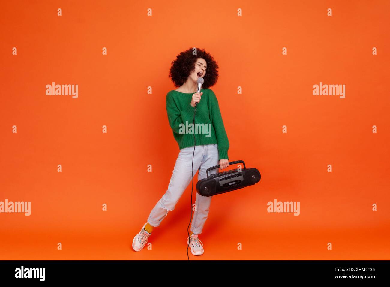 Full length portrait of woman with Afro hairstyle wearing green casual style sweater standing with tape recorder and microphone, singing songs. Indoor studio shot isolated on orange background. Stock Photo